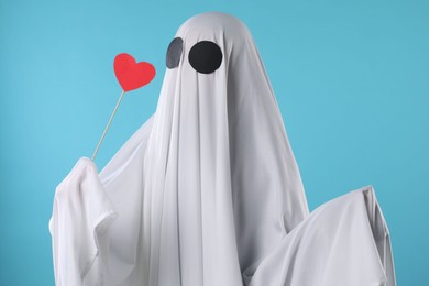 Cute ghost. Person covered with white sheet holding stick with red heart on light blue background