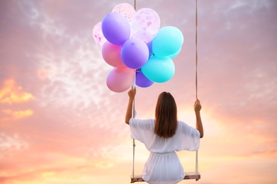 Dream world. Young woman with bright balloons swinging, sunset sky on background