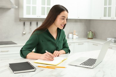 Photo of Home workplace. Happy woman with pen and notebook working on laptop at marble desk in kitchen