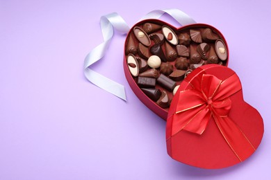 Heart shaped box with delicious chocolate candies and ribbon on lilac background, flat lay. Space for text