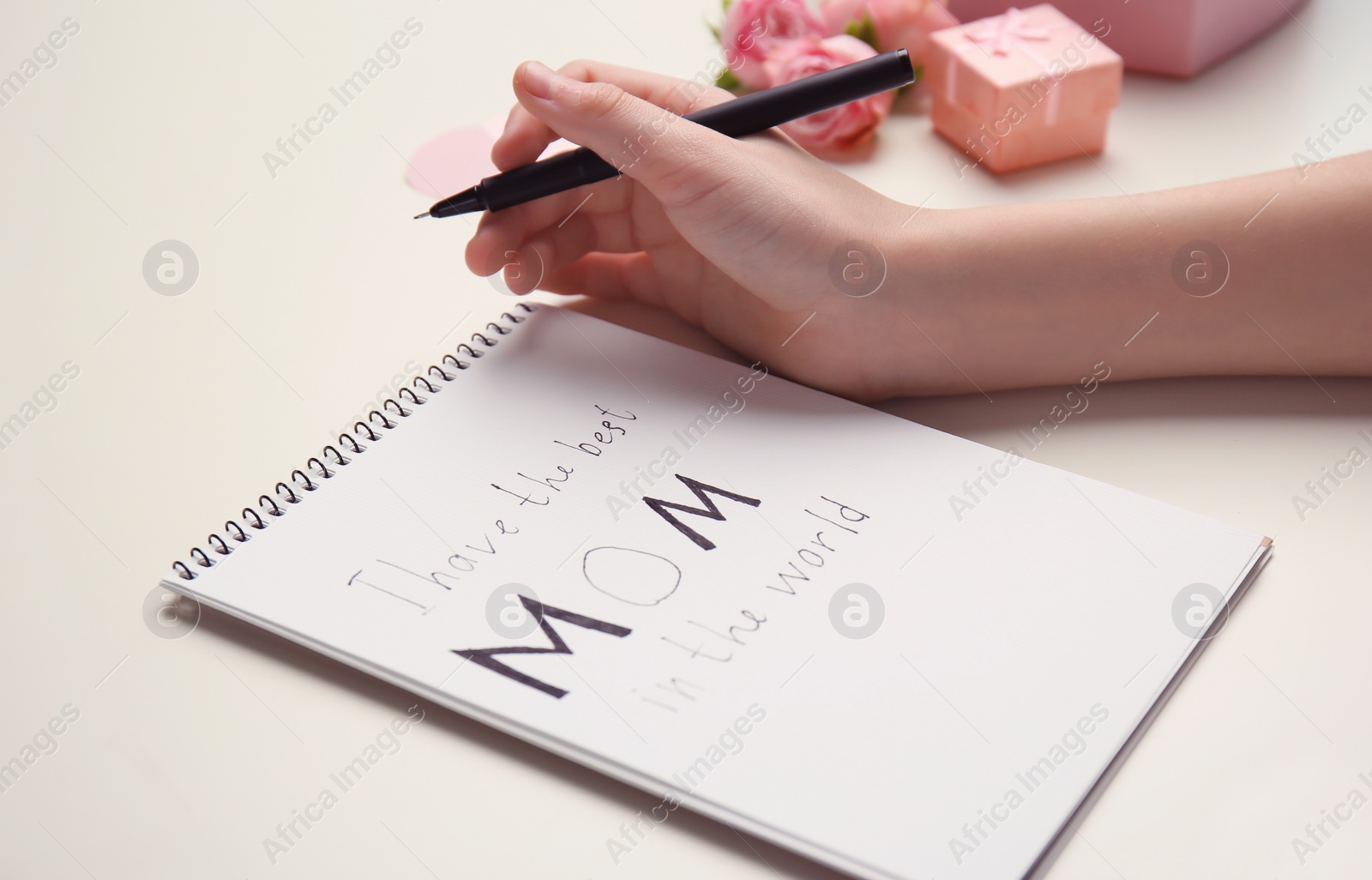 Photo of Little child writing congratulation for Mother's Day in notebook on light background