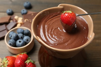 Photo of Dipping fresh strawberry in fondue pot with melted chocolate at wooden table, closeup