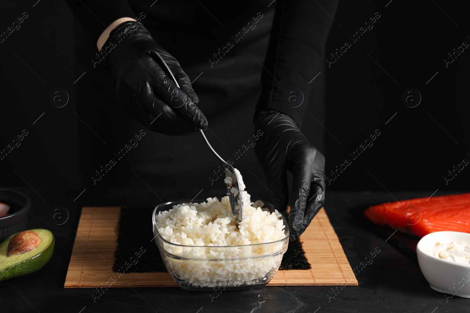 Photo of Chef in gloves taking cooked rice for sushi with spoon at dark table, closeup