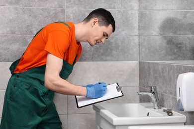 Photo of Young plumber writing results of examining sink in bathroom