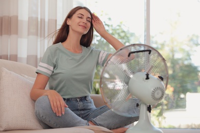 Woman enjoying air flow from fan on sofa in living room. Summer heat