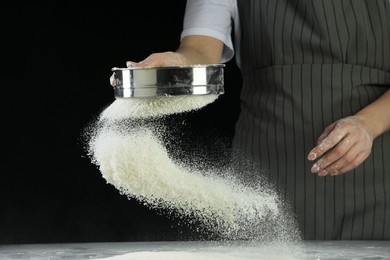 Photo of Woman sieving flour at table against black background, closeup