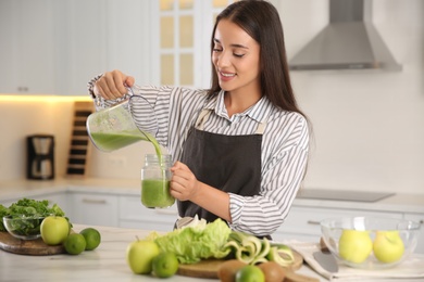 Young woman pouring fresh green juice into mason jar at table in kitchen