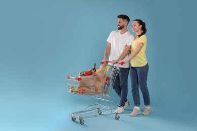 Photo of Happy couple with shopping cart full of groceries on light blue background