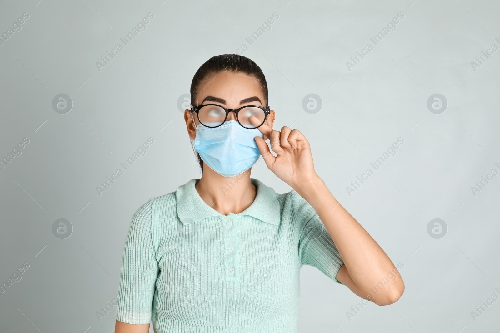 Photo of Woman wiping foggy glasses caused by wearing medical mask on light background