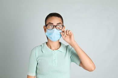 Woman wiping foggy glasses caused by wearing medical mask on light background