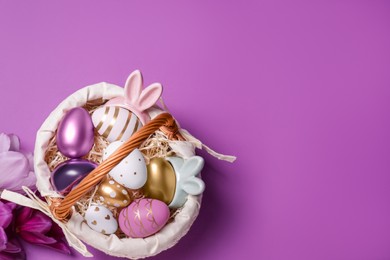Photo of Easter basket with painted eggs and flowers on purple background, flat lay. Space for text