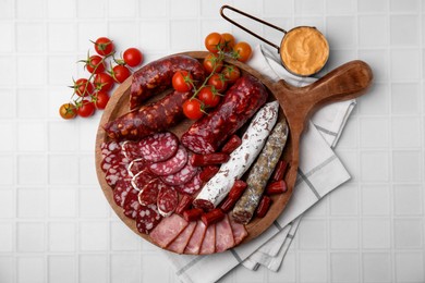 Different types of delicious sausages, sauce and tomatoes on white tiled table, flat lay