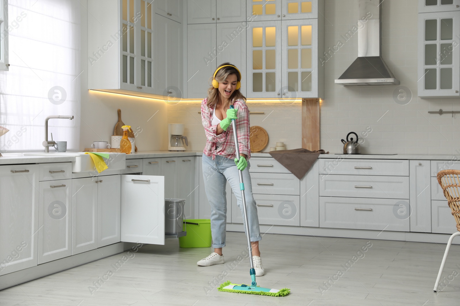 Photo of Beautiful young woman with headphones singing while cleaning kitchen