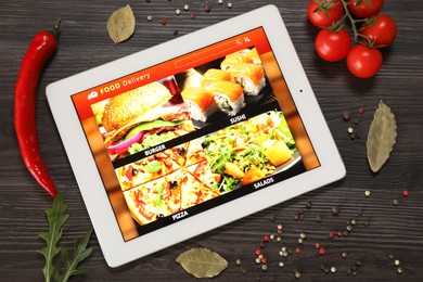 Photo of Modern tablet with open page for online food ordering surrounded by scattered products on wooden table, flat lay. Delivery service