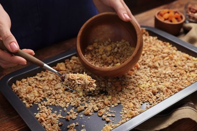 Photo of Woman putting granola from baking tray into bowl at table, closeup