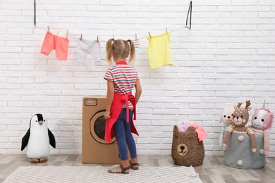 Photo of Little girl playing with toy cardboard washing machine indoors