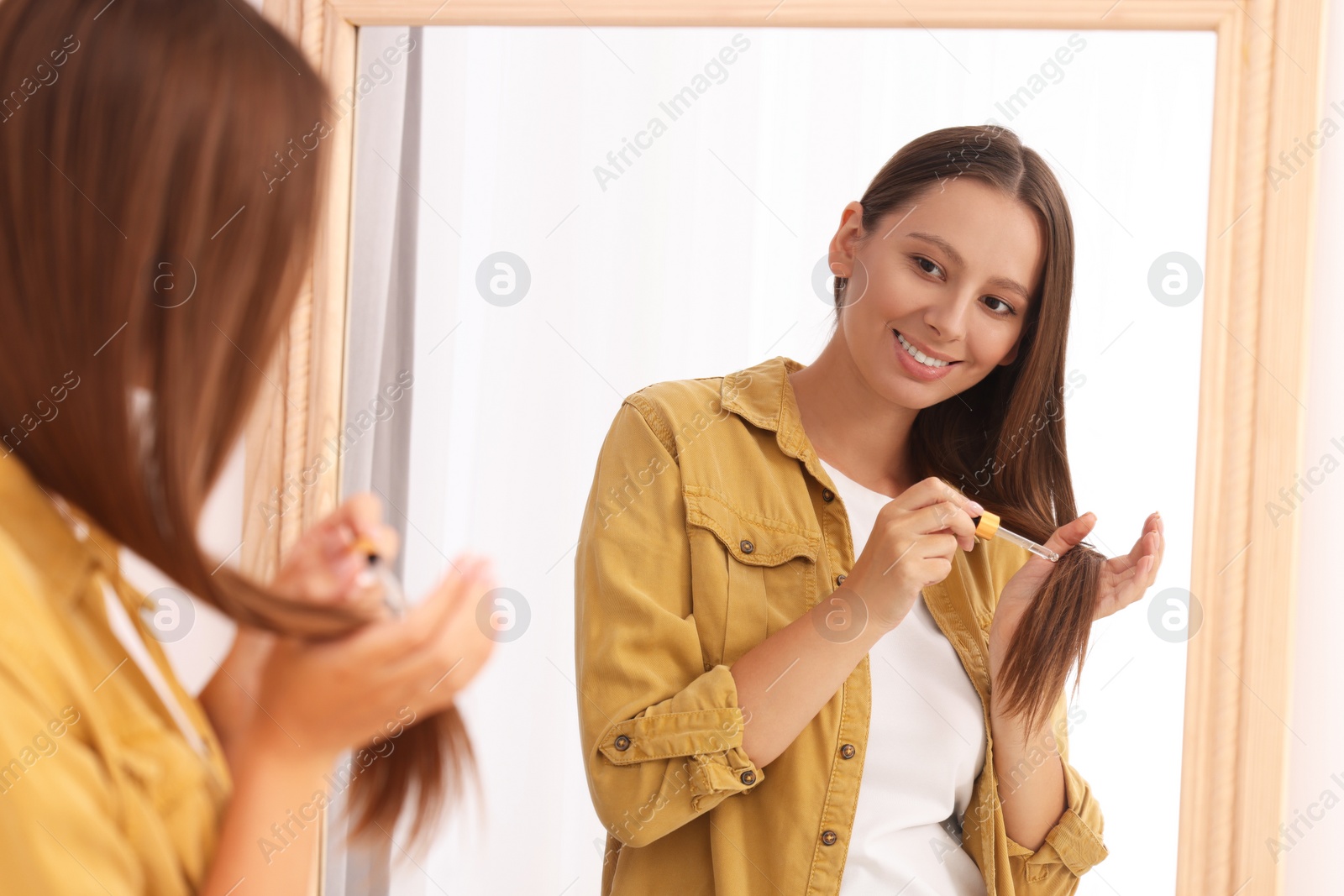 Photo of Beautiful woman applying serum onto hair near mirror indoors