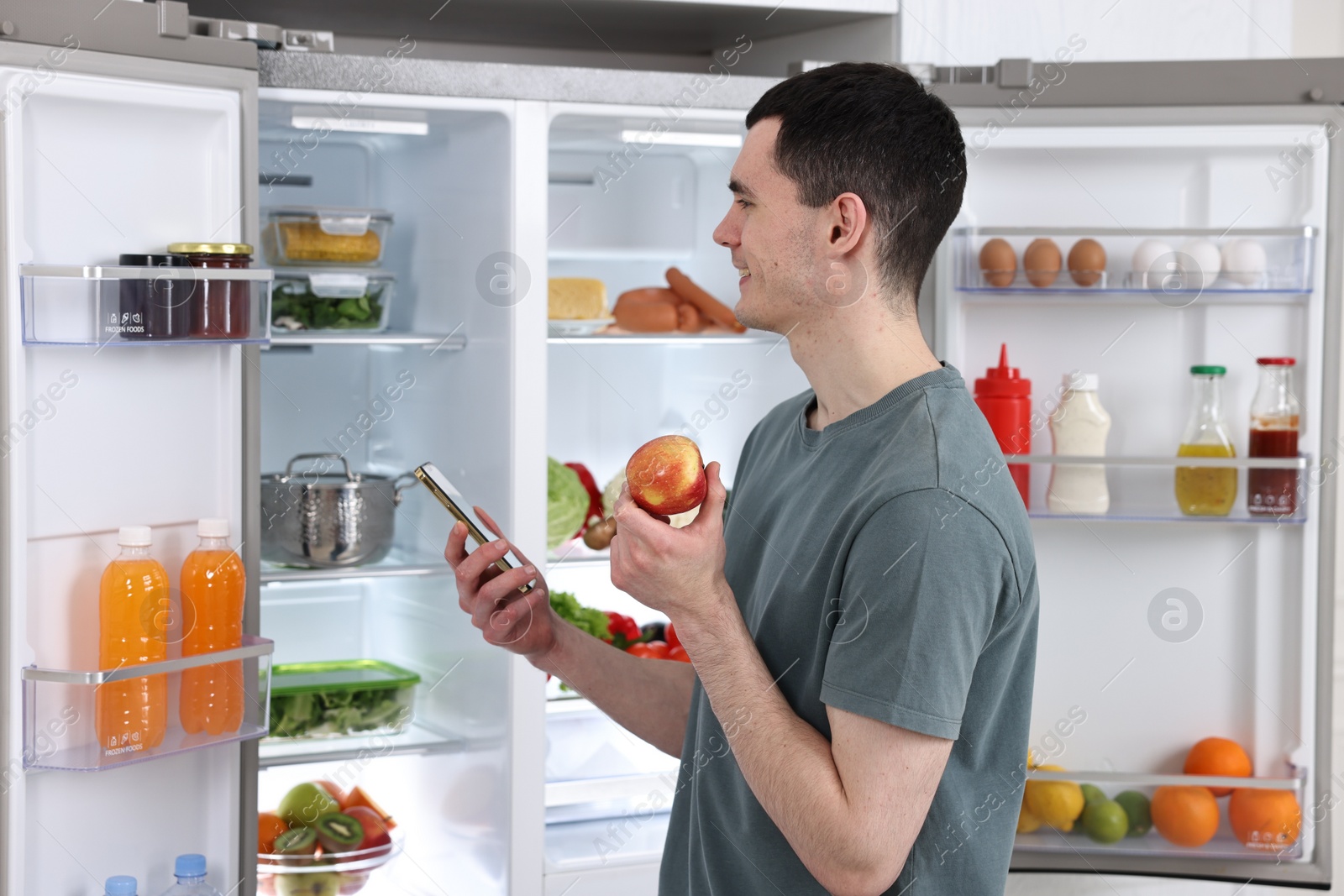 Photo of Happy man with apple and smartphone near refrigerator in kitchen
