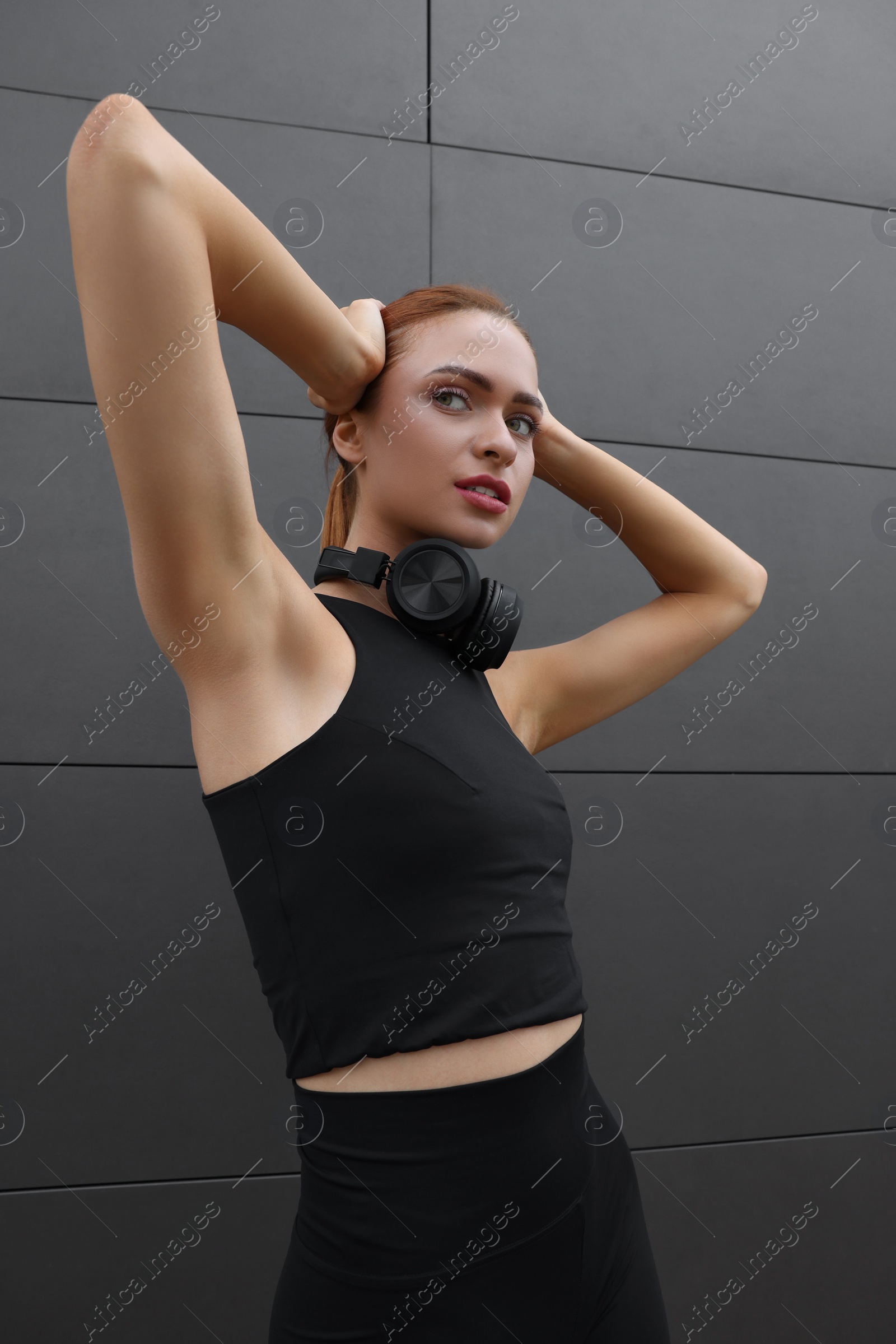 Photo of Beautiful woman in stylish gym clothes with headphones posing near dark grey wall on street