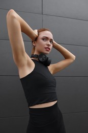Photo of Beautiful woman in stylish gym clothes with headphones posing near dark grey wall on street
