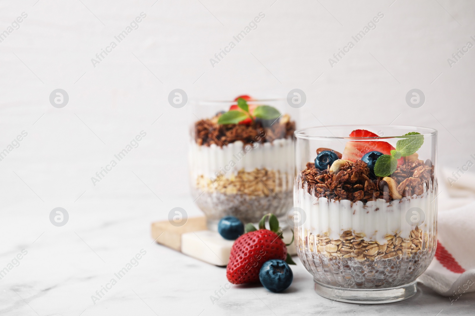 Photo of Tasty granola with berries, nuts, yogurt and chia seeds in glasses on white marble table, space for text