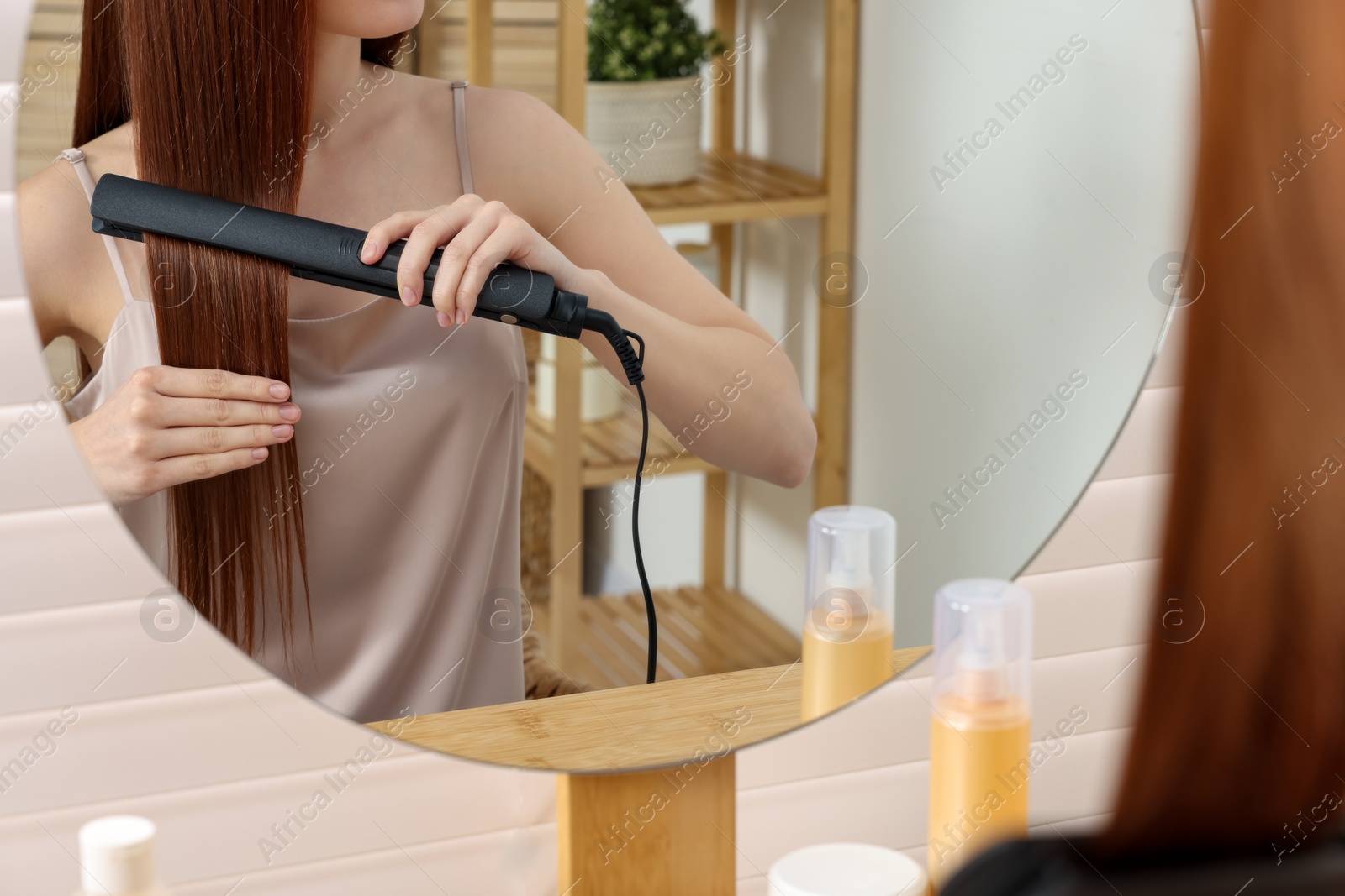 Photo of Beautiful woman using hair iron near mirror in room, closeup