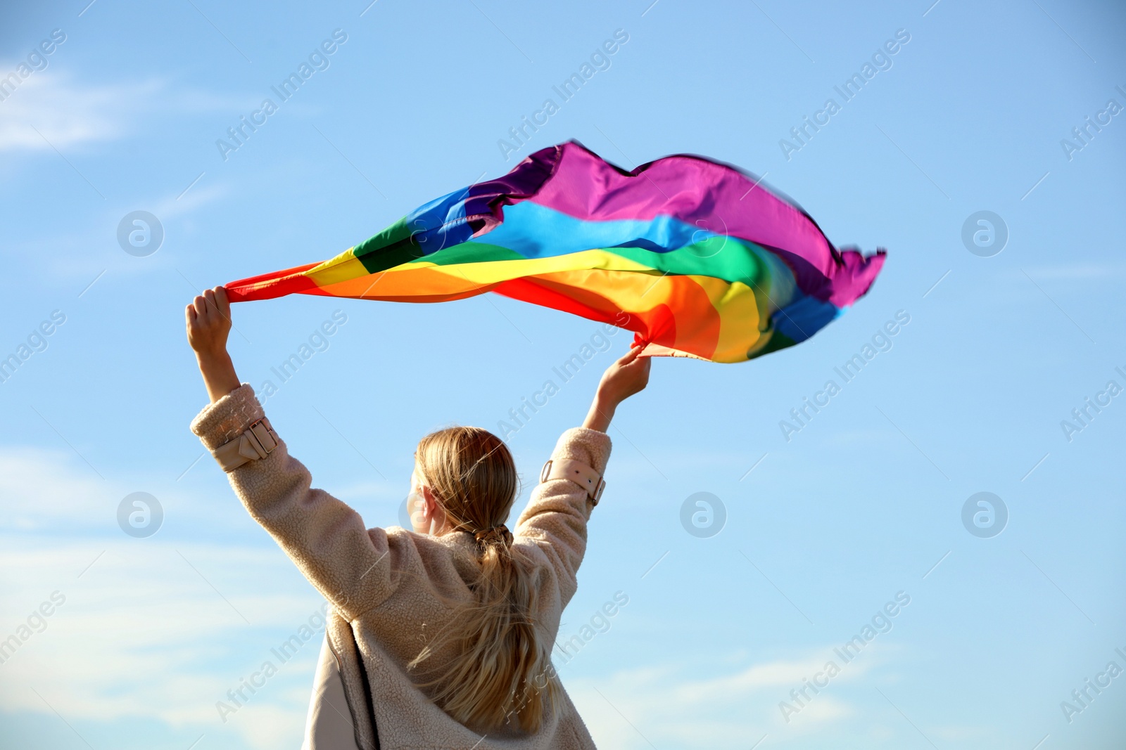 Photo of Woman holding bright LGBT flag against blue sky, back view