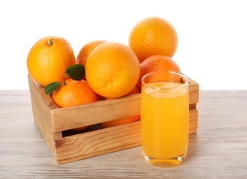 Fresh oranges in crate and glass of juice on light wooden table against white background