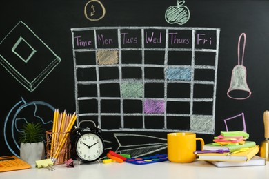 Photo of Different stationery, alarm clock and cup on white table near blackboard with drawn school timetable