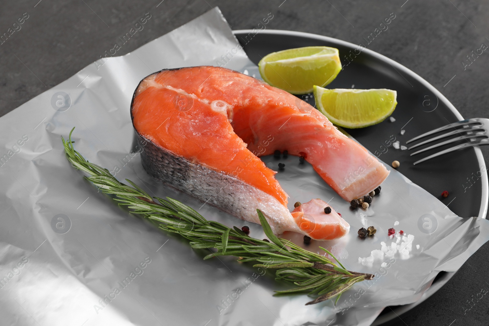 Photo of Aluminum foil with raw fish, lime, rosemary and spices on grey table, closeup. Baking salmon