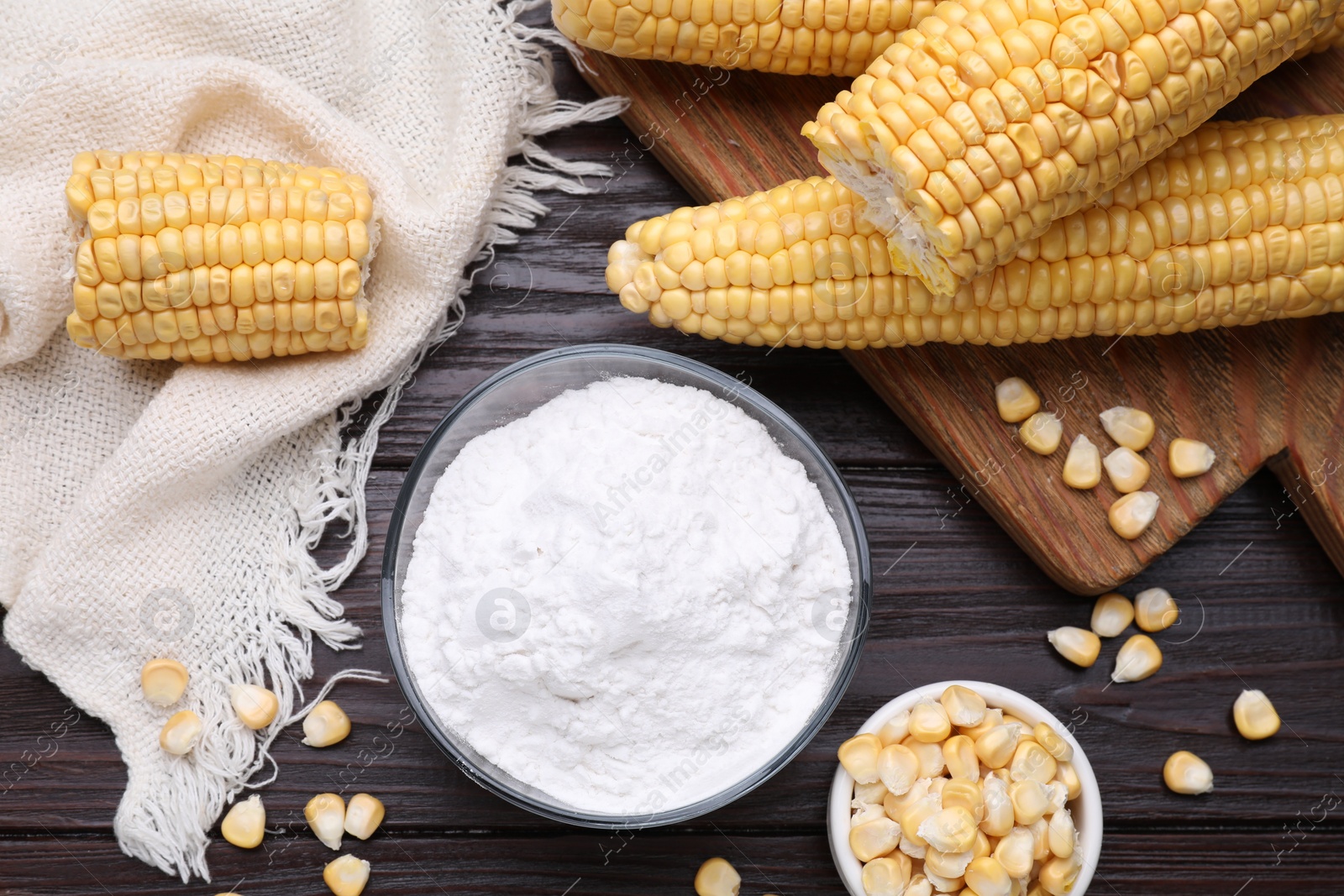 Photo of Bowl with corn starch, ripe cobs and kernels on dark wooden table, flat lay