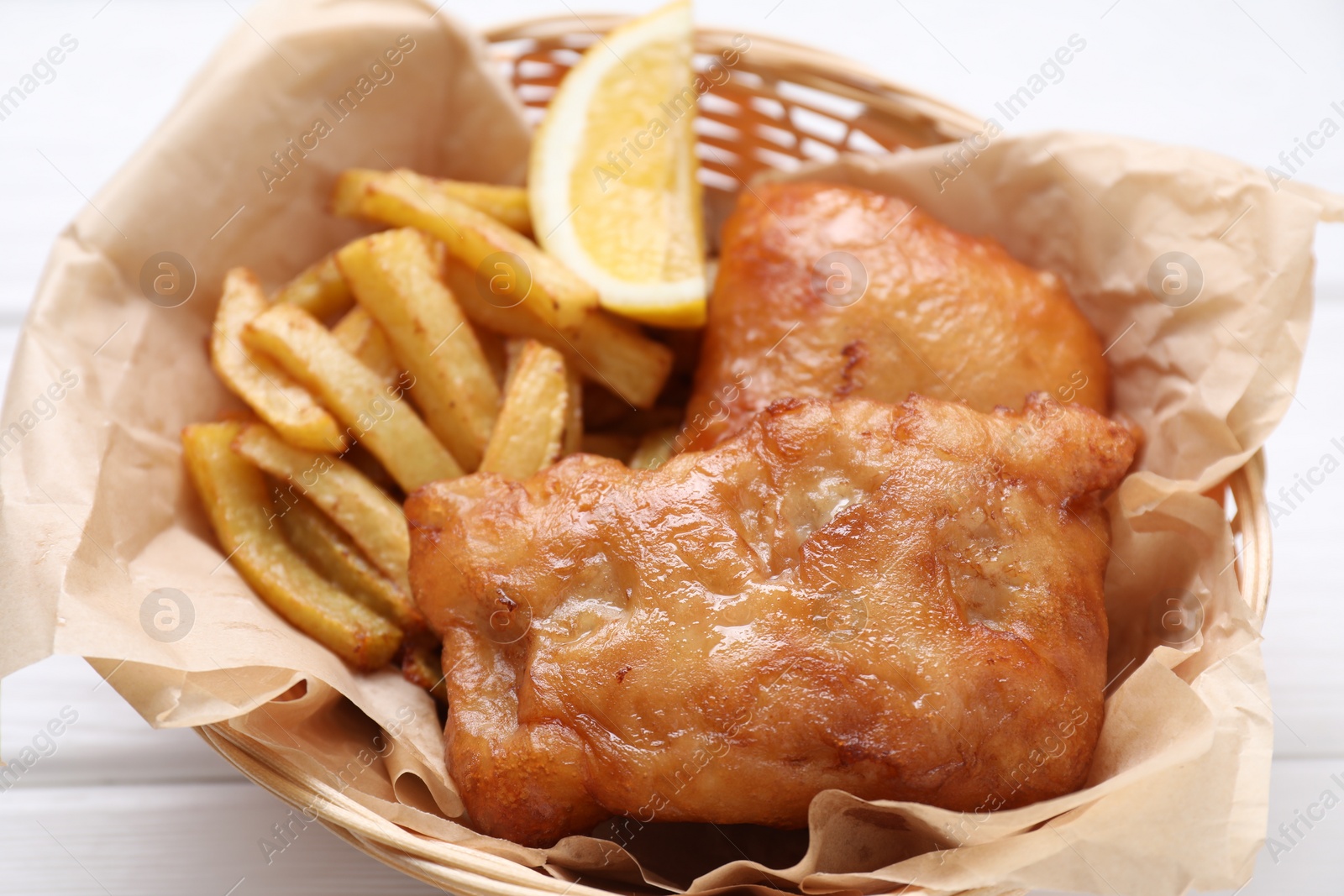 Photo of Tasty fish, chips and lemon in wicker bowl on white wooden table, closeup