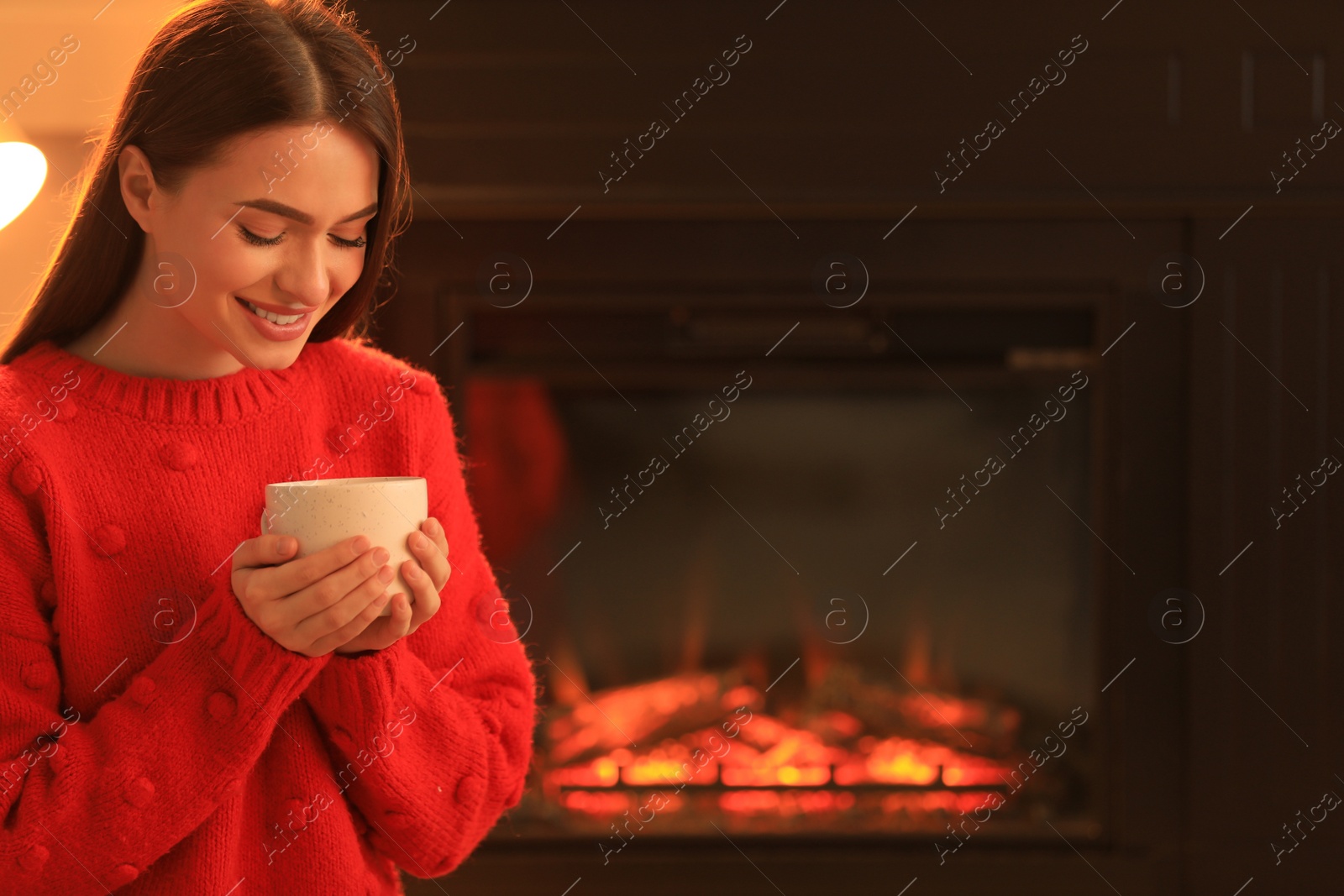 Photo of Young woman with cup of hot drink near fireplace indoors. Cozy atmosphere