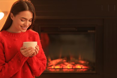 Young woman with cup of hot drink near fireplace indoors. Cozy atmosphere