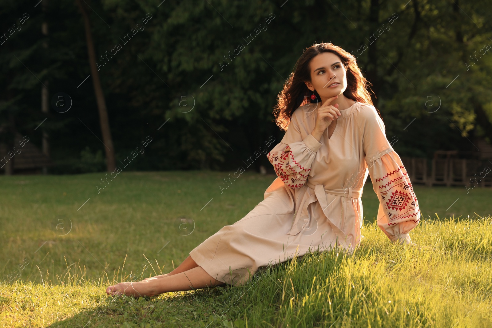 Photo of Beautiful woman in embroidered dress sitting on green grass outdoors, space for text. Ukrainian national clothes