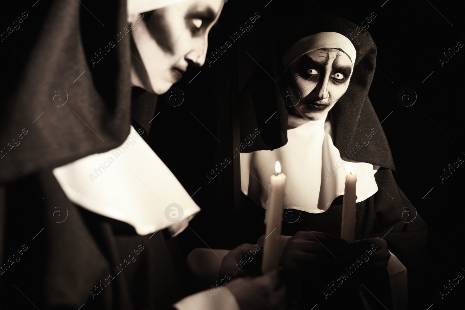 Photo of Scary devilish nun with burning candle near mirror on black background. Halloween party look