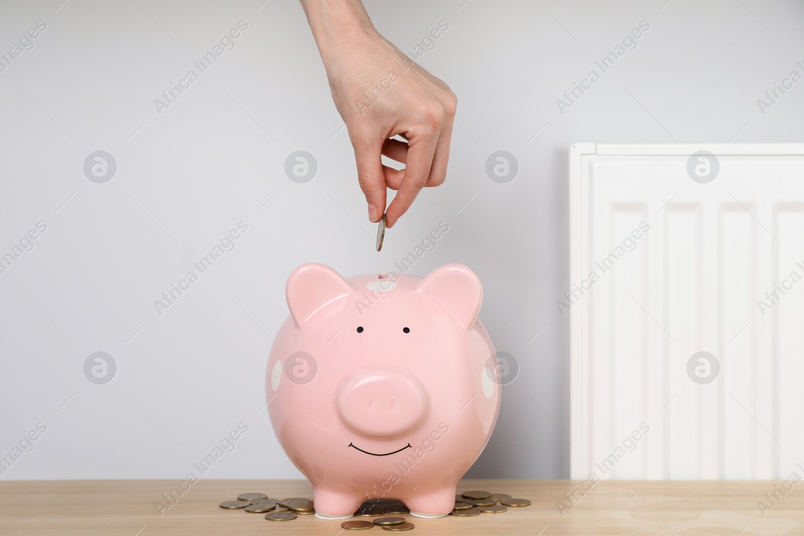 Photo of Woman putting coin into piggy bank near heating radiator, closeup