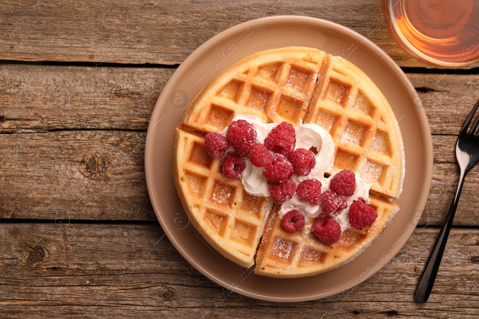 Photo of Tasty Belgian waffle with fresh raspberries, whipped cream, cup of tea and fork on wooden table, flat lay