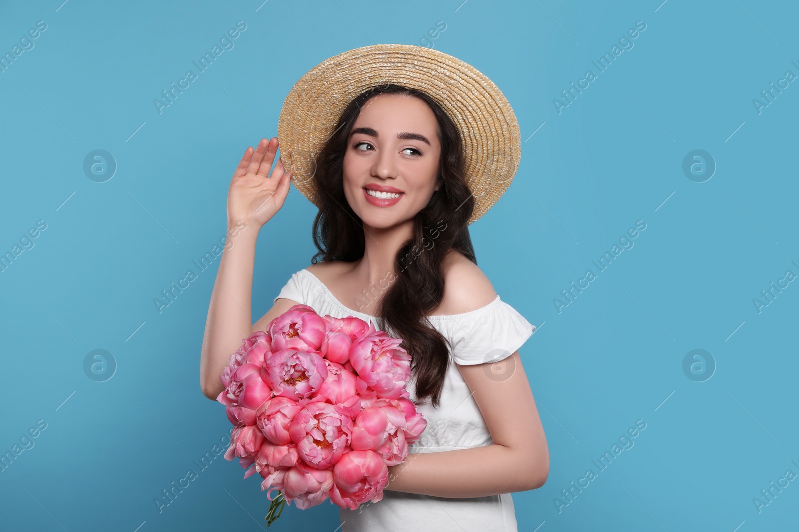 Photo of Beautiful young woman in straw hat with bouquet of pink peonies against light blue background