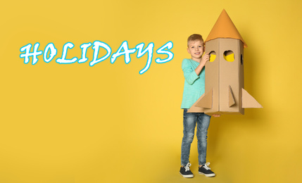 Image of School holidays. Cute little child playing with cardboard rocket near yellow wall. Banner design 