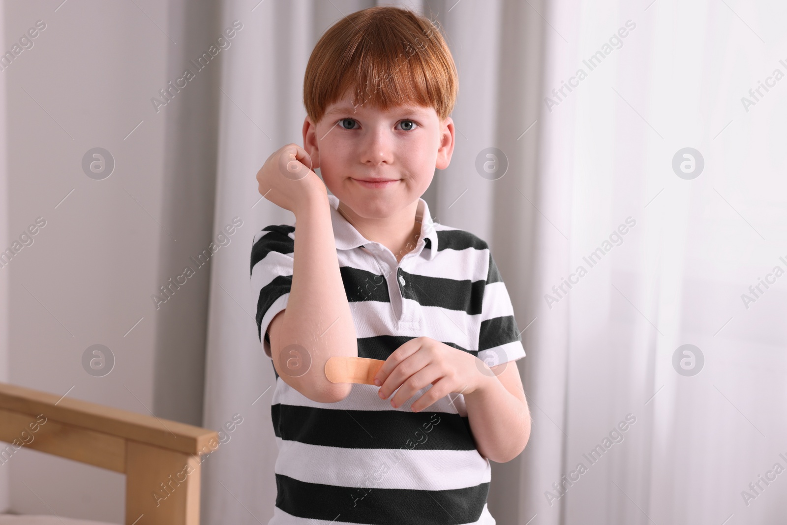 Photo of Little boy putting sticking plaster onto elbow indoors