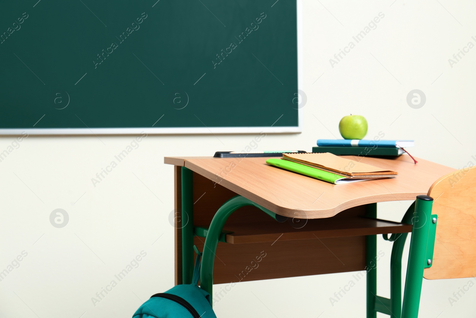 Photo of Wooden school desk with stationery, apple and backpack near chalkboard in classroom