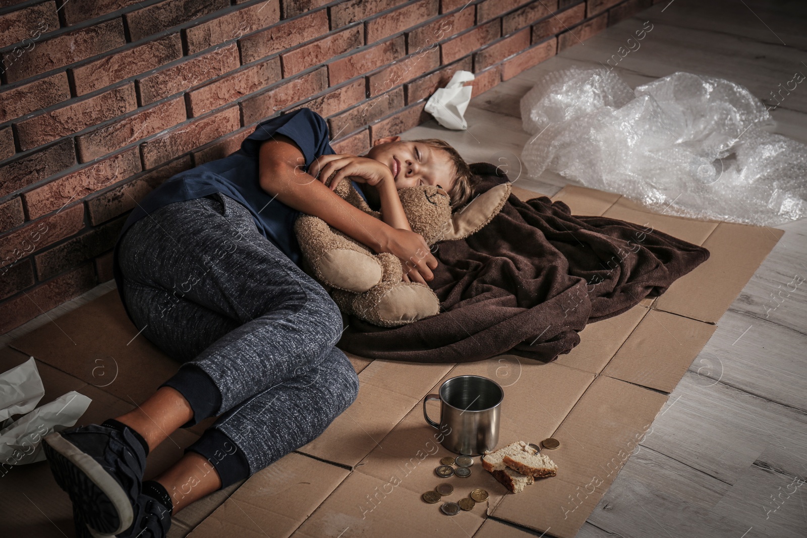 Photo of Poor homeless boy sleeping with his toy on floor near brick wall