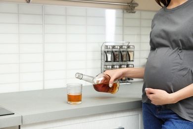 Photo of Future mother pouring alcohol drink from bottle into glass in kitchen, closeup. Bad habits during pregnancy