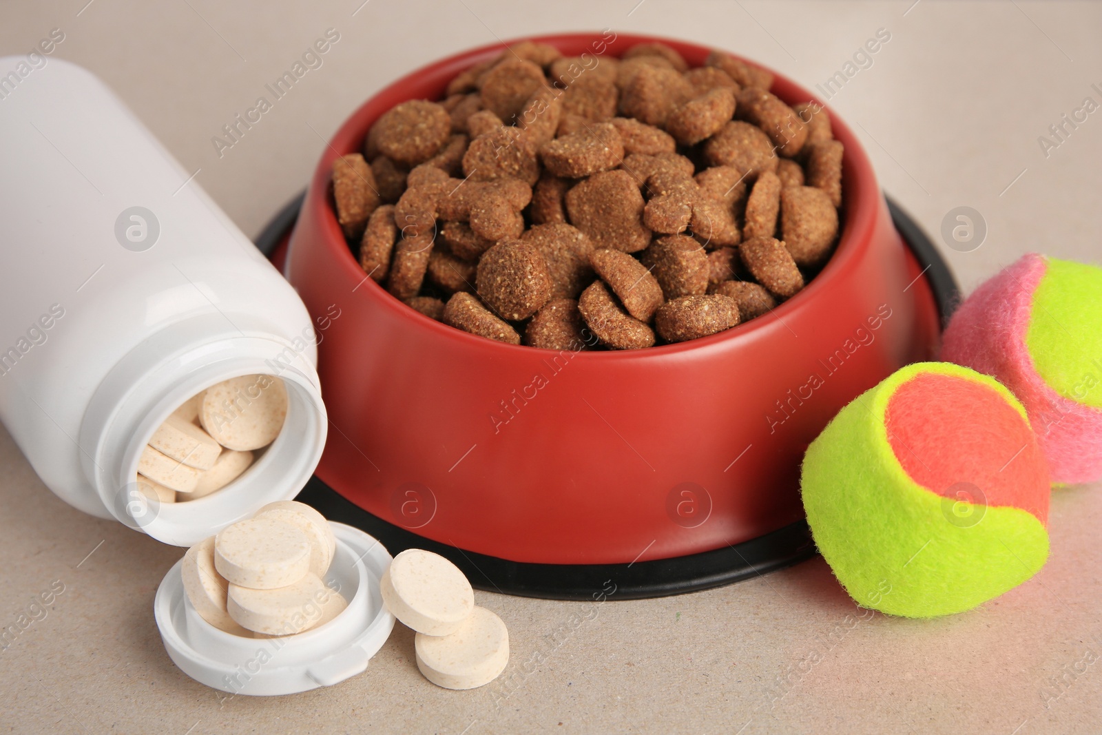 Photo of Bottle with vitamins, dry pet food in bowl and toy balls on beige background, closeup