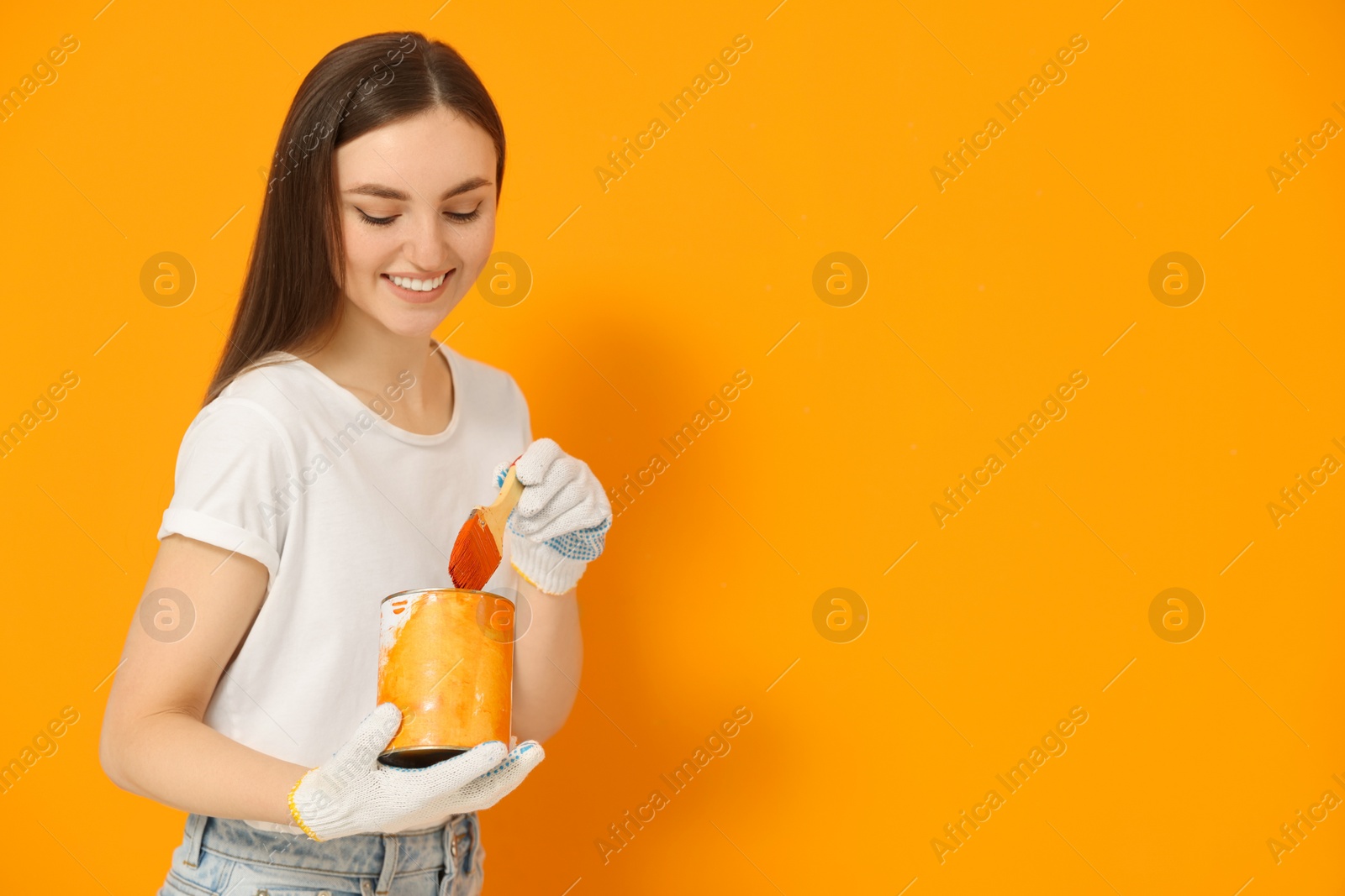 Photo of Happy designer with brush taking paint from can near orange wall, space for text