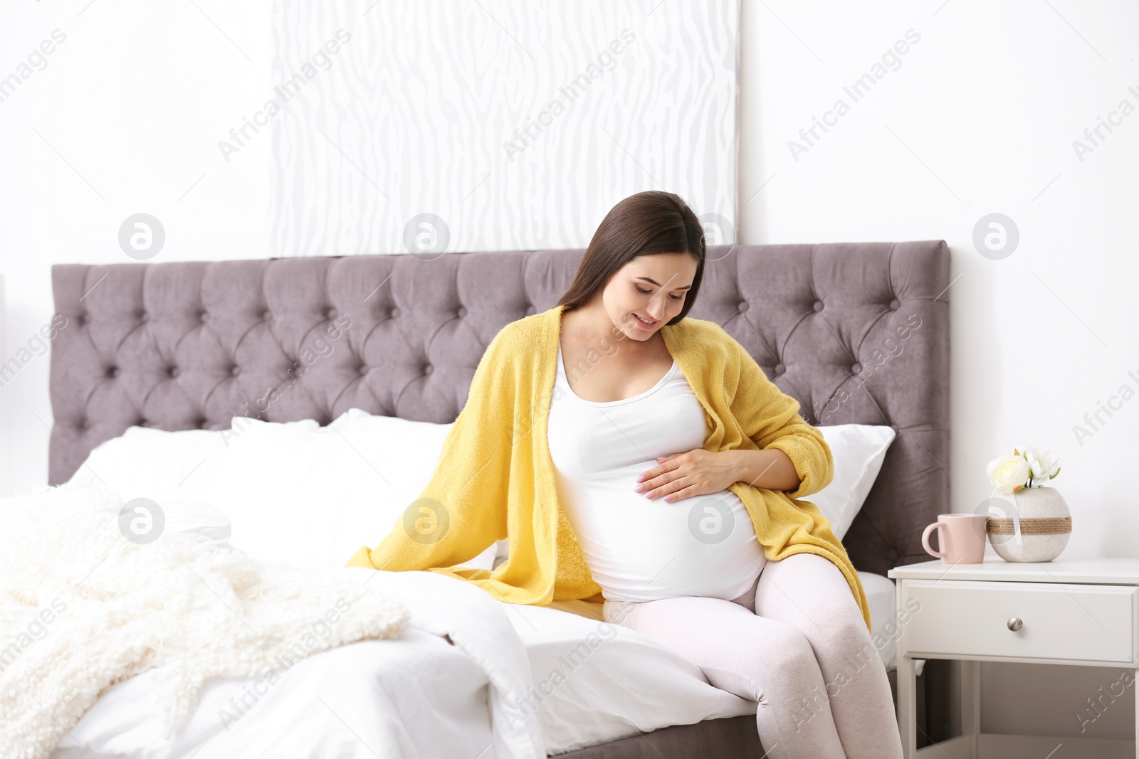 Photo of Young beautiful pregnant woman sitting on bed and touching her belly at home