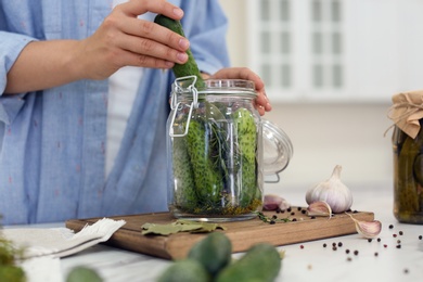 Photo of Woman putting cucumber into pickling jar at table in kitchen, closeup