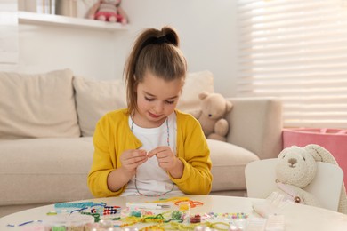 Cute little girl making beaded jewelry at table in room