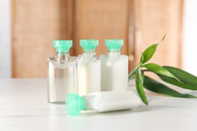 Photo of Mini bottles of cosmetic products and green branch on white table against blurred background